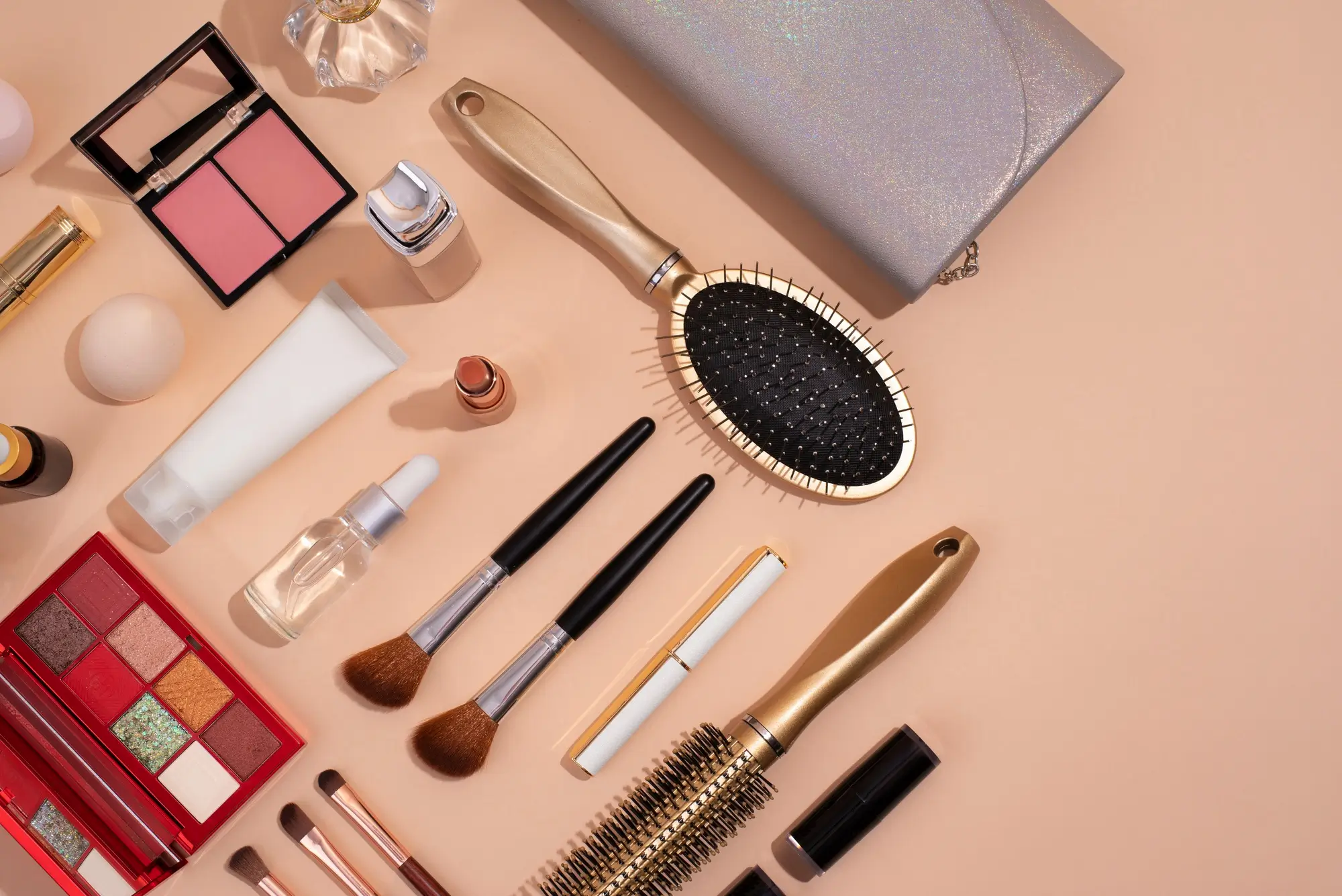 Flat lay of makeup brushes, hairbrushes, lipstick, foundation bottles, and styling tools arranged on a peach background at a beauty and hair salon in Denver.